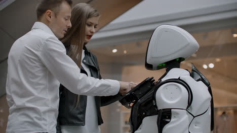 Ein-Mann-Und-Eine-Frau-Im-Einkaufszentrum-Interagieren-Mit-Einem-Roboterberater,-Indem-Sie-Auf-Den-Bildschirm-Tippen-Und-Lächeln.-Cyborg-Android-Hilft-Menschen